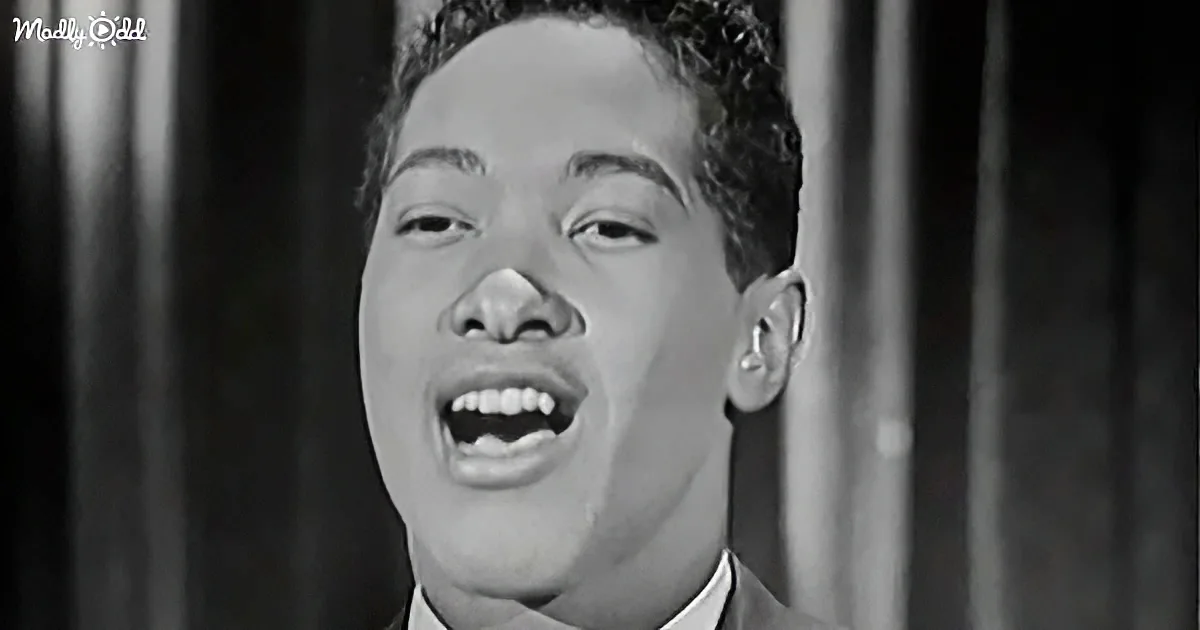 A man in a suit sings passionately, mouth open, against a dark curtain backdrop.