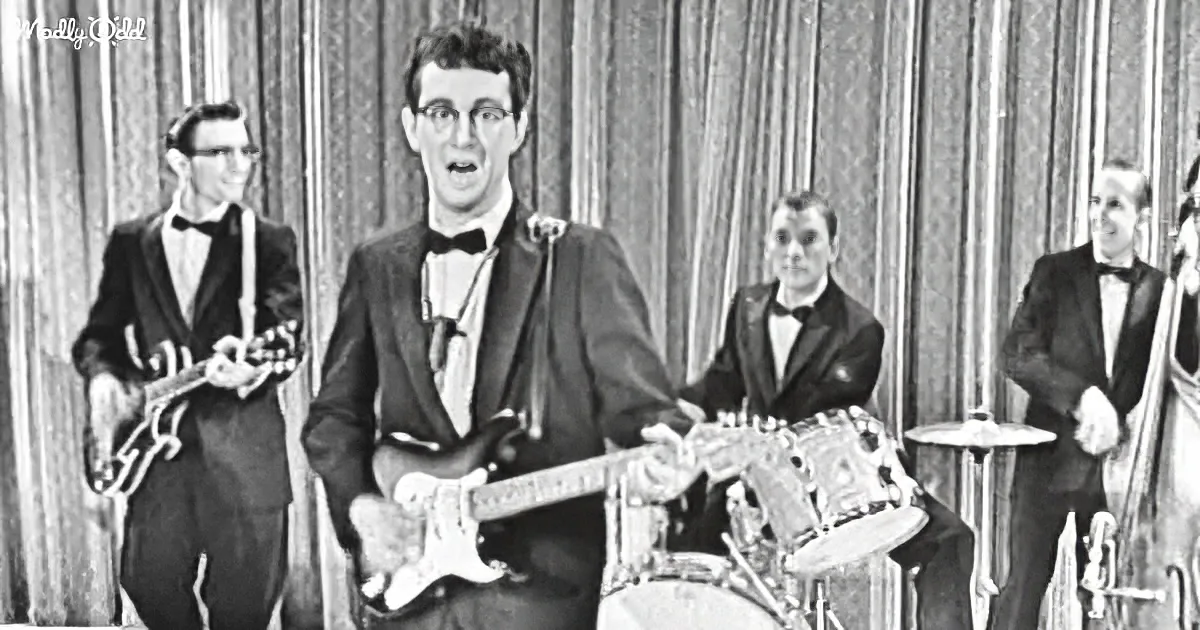 A man in a tuxedo plays a white electric guitar on stage with a band.