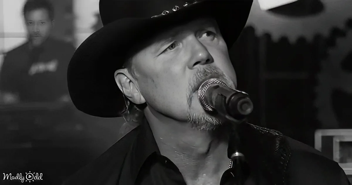 A man in a black hat sings into a microphone, wearing a dark shirt, with another person in the background.