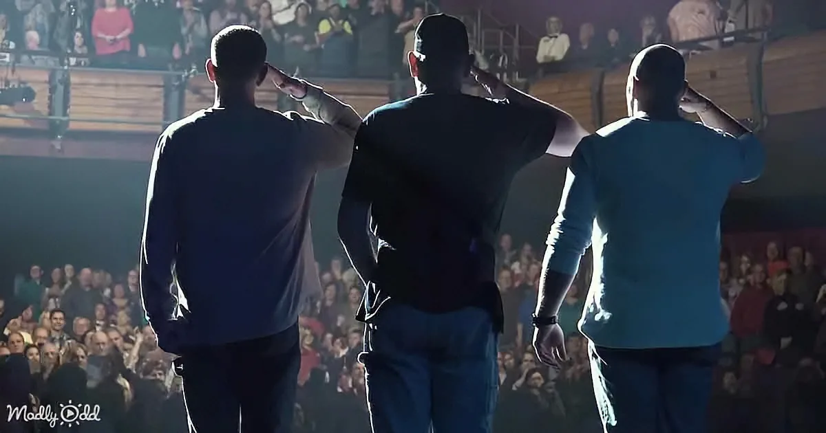 Three men in a line, backs to camera, saluting in front of a large audience.