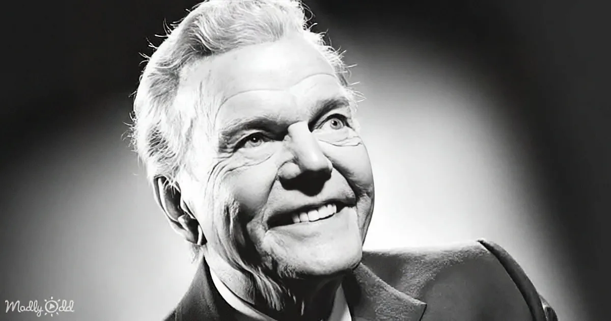 An older man with white hair, wearing a suit, smiles warmly under a spotlight.