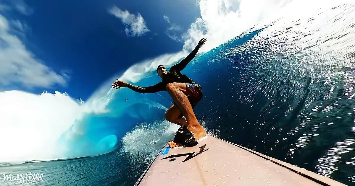A surfer rides a wave, arms outstretched, on a sunny day with a vibrant blue sky.