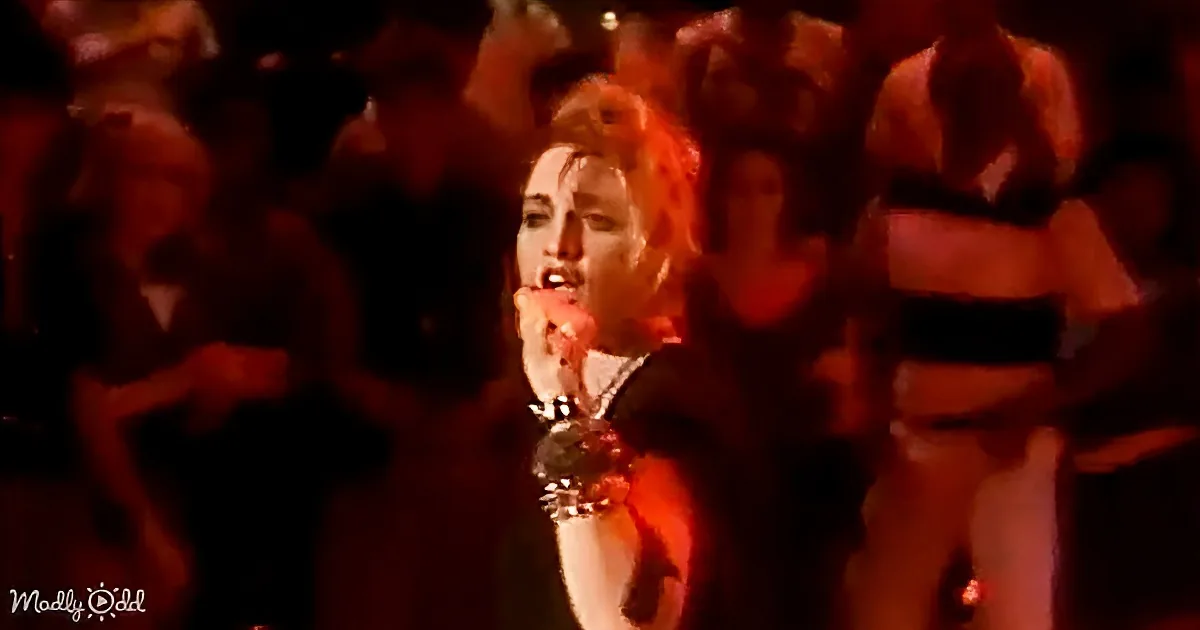 A person with red hair and a microphone, surrounded by dancers in a dimly lit setting.