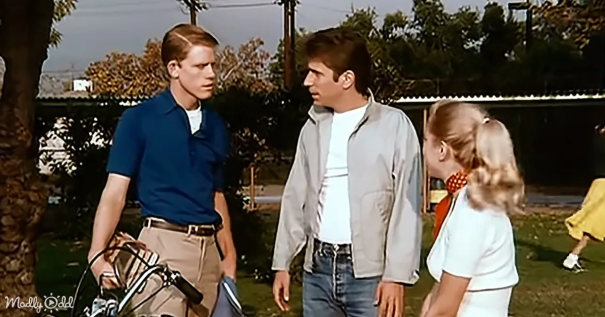 A man in a blue shirt and khaki pants stands with a bicycle, talking to another man in a gray jacket and jeans, while a...