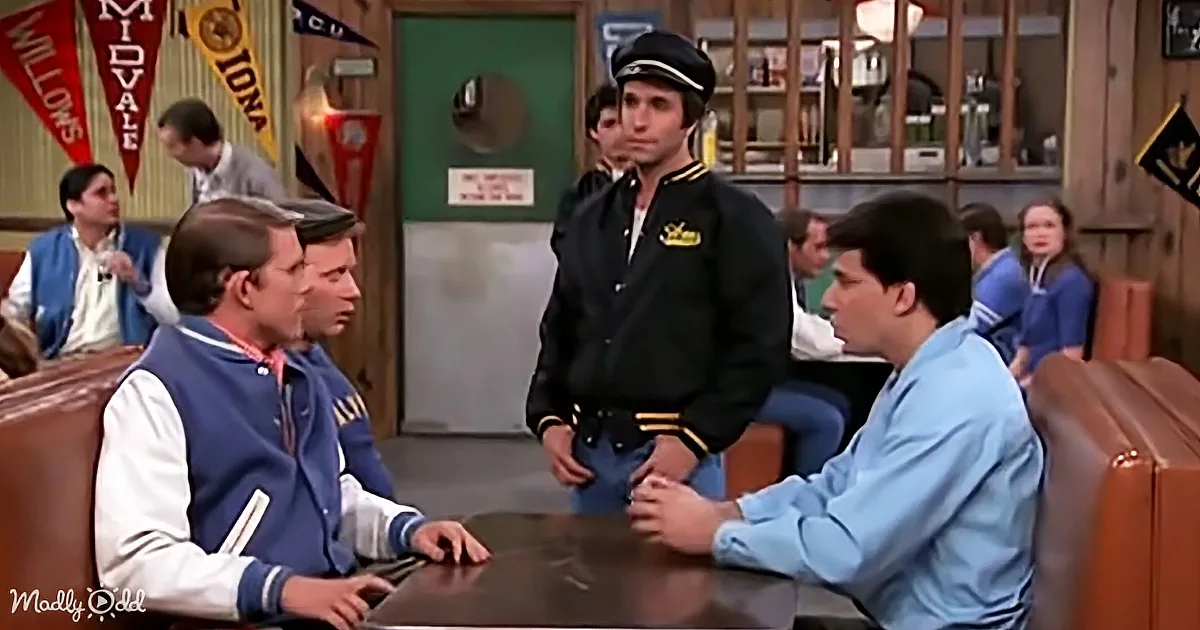 A man in a varsity jacket and another in a uniform are seated at a table, engaged in conversation.