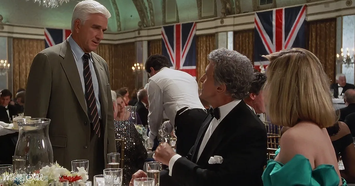 A man in a suit stands at a formal event with British flags in the background, surrounded by other guests in elegant attire.