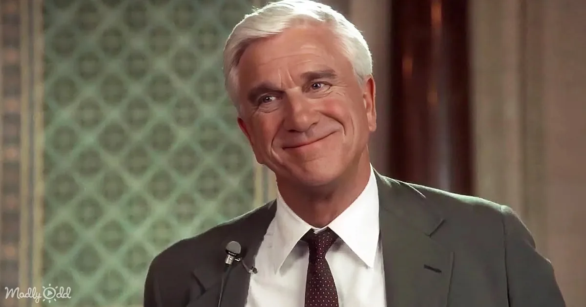 A man in a suit and tie smiles while speaking into a microphone, with a patterned background.