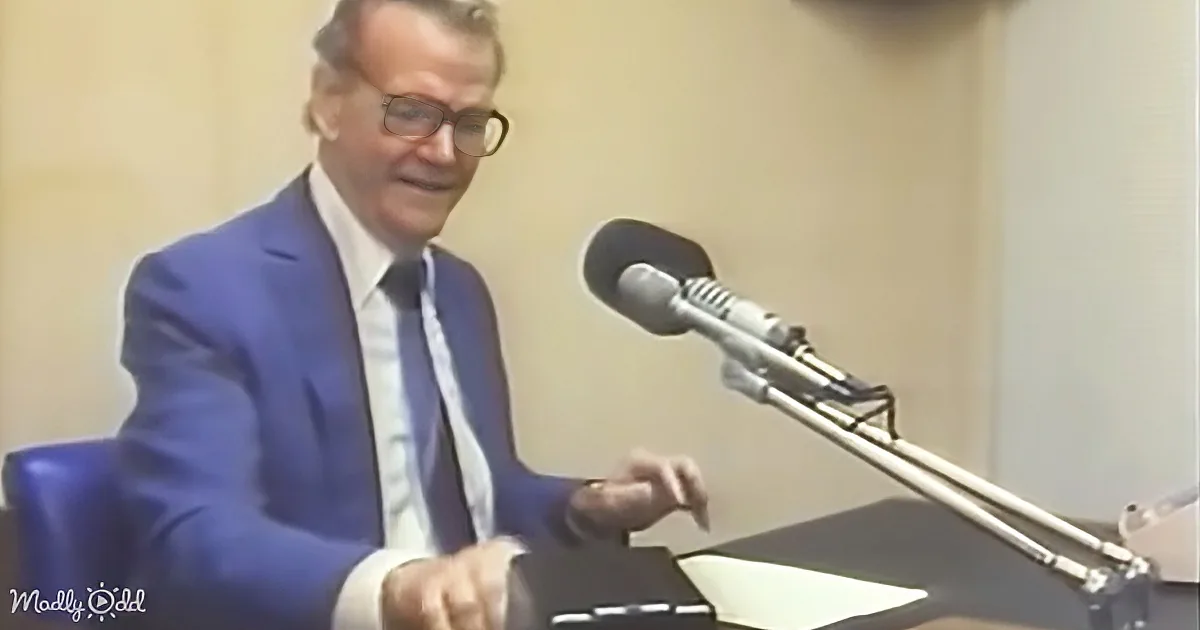 A man in a blue suit sits at a desk with a microphone, smiling and gesturing.
