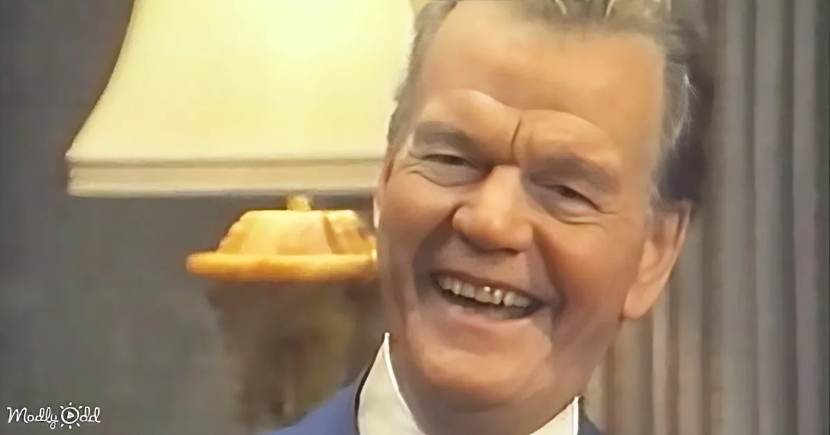 A smiling man in a suit and tie, with a lamp and curtain in the background.