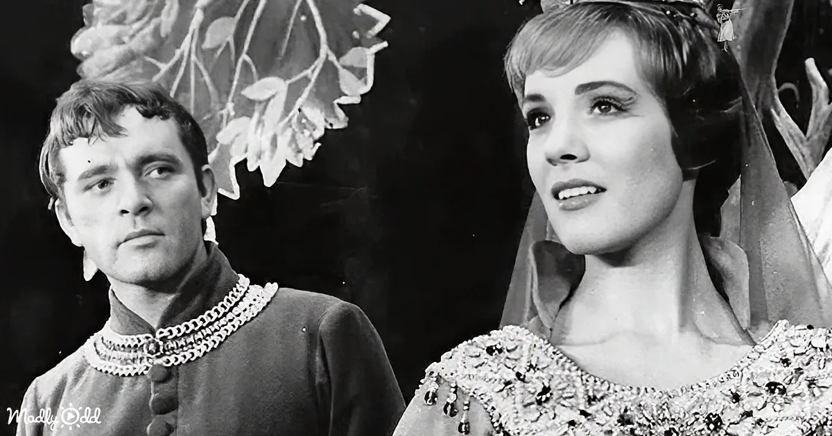 A woman in a regal dress and crown stands beside a man in a medieval outfit, both in a theatrical setting.