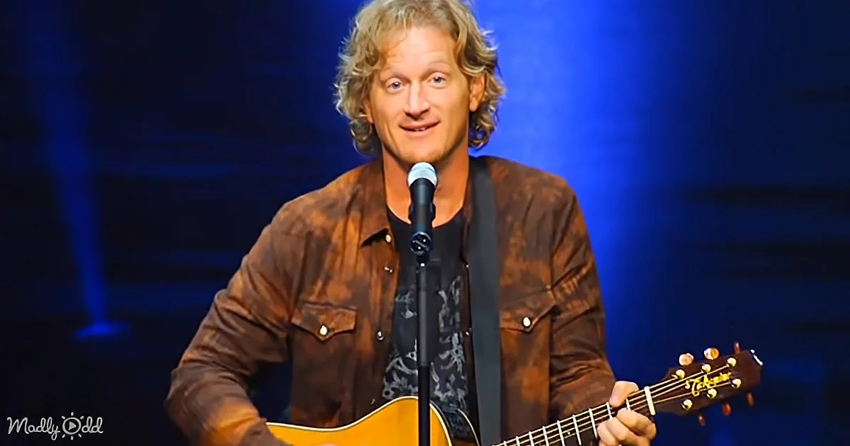 A man with blonde hair, wearing a brown jacket, plays an acoustic guitar on stage with a microphone in front of him.