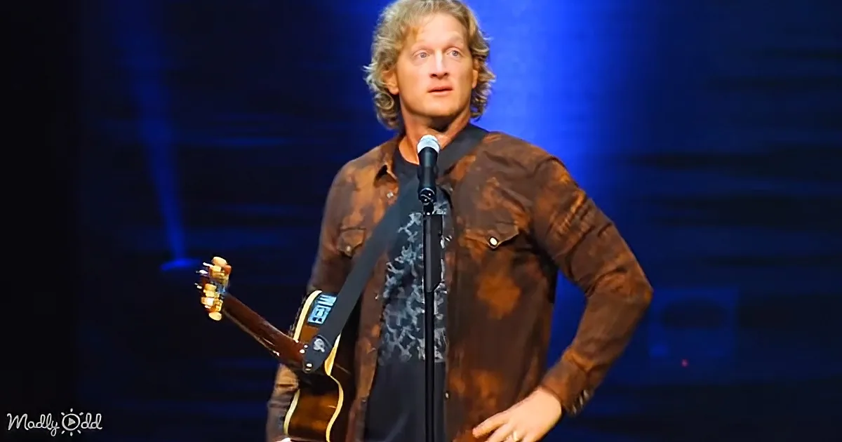 A man with curly hair stands on stage, holding a guitar, wearing a brown jacket and black shirt, with a microphone in...