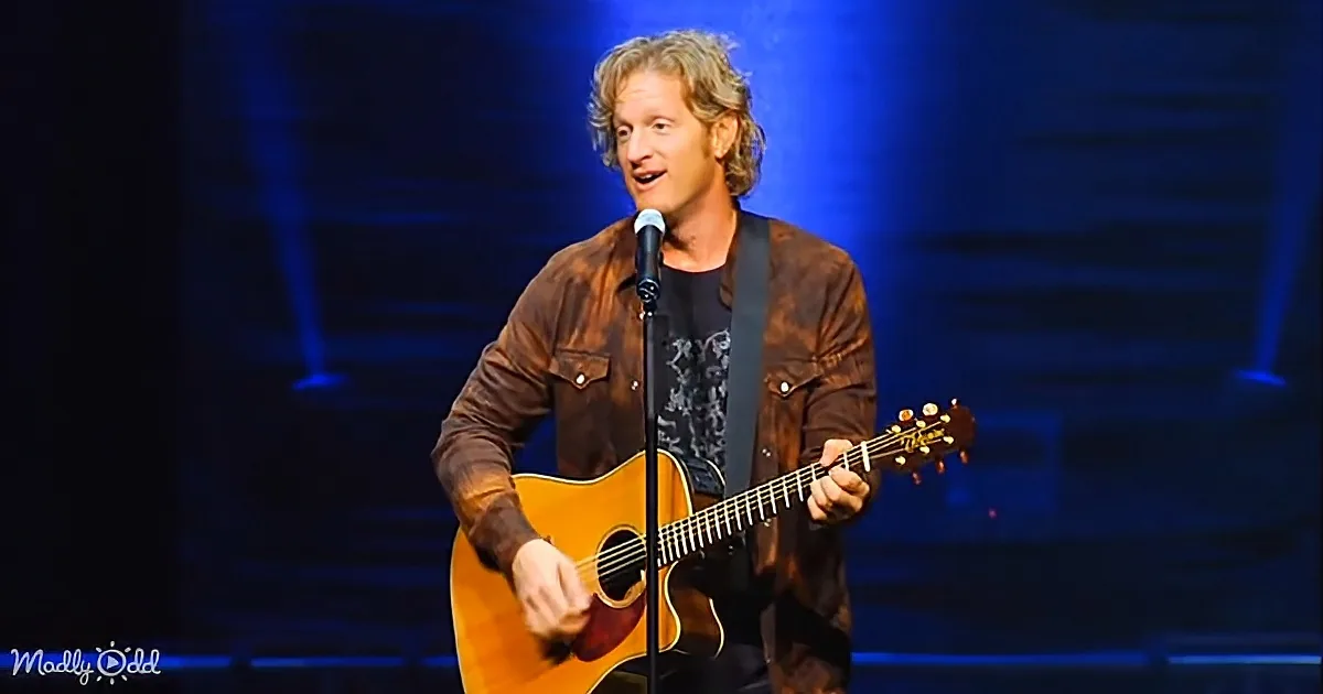 A man with long hair, wearing a brown jacket, plays an acoustic guitar on stage, singing into a microphone.