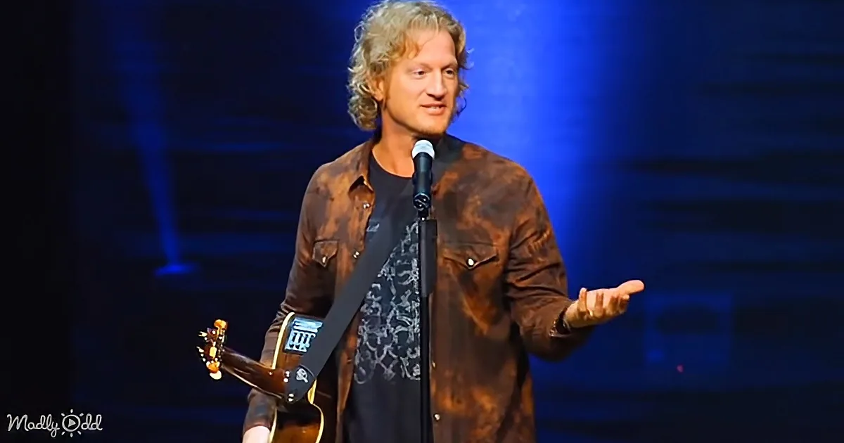 A man with long hair, wearing a brown jacket and black shirt, holds a guitar while speaking into a microphone on stage.