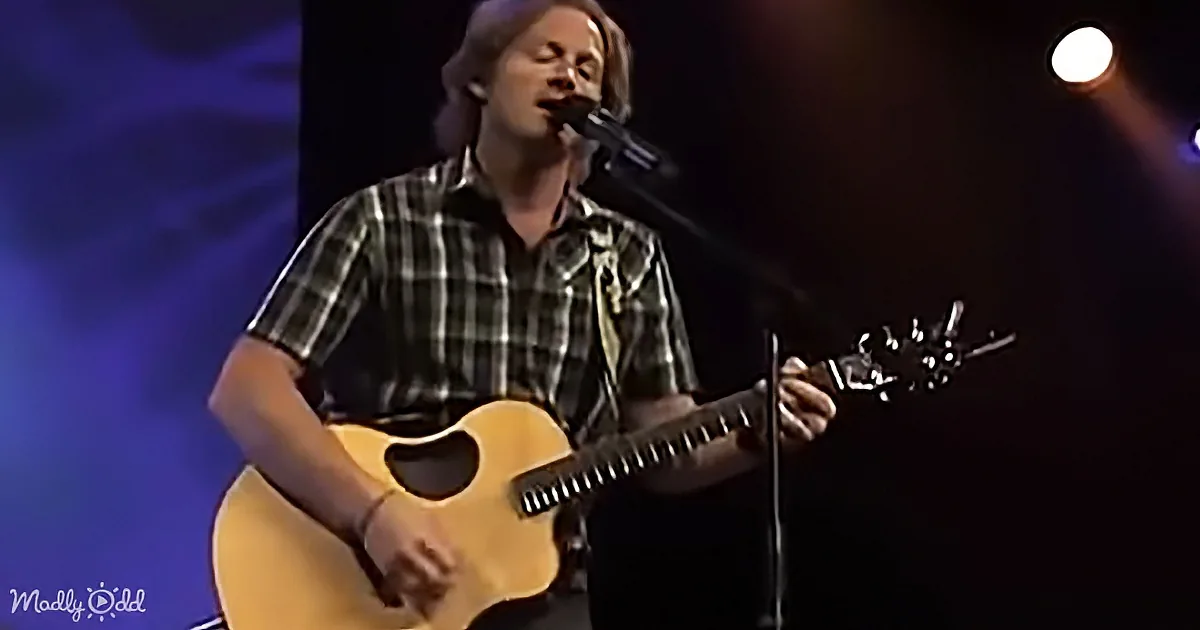A man in a plaid shirt plays an acoustic guitar and sings into a microphone on stage.