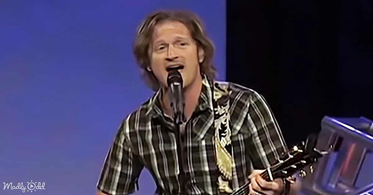 A man in a plaid shirt sings into a microphone while holding a guitar.