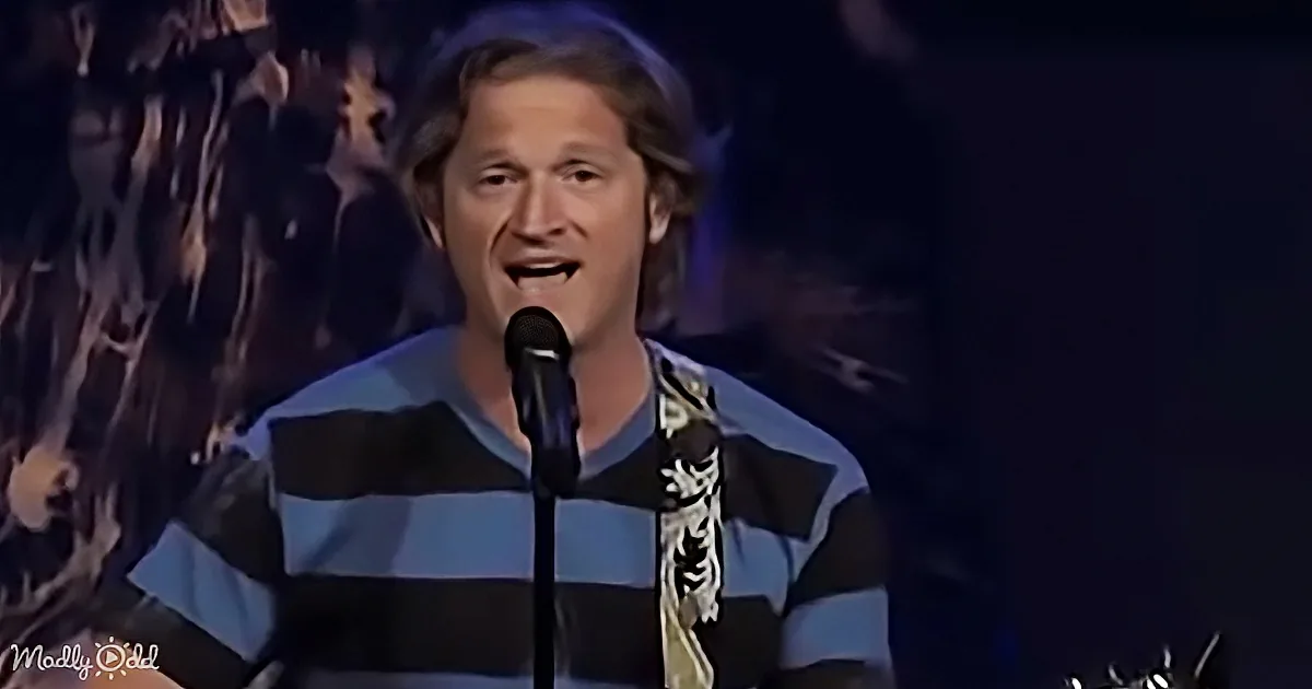 A man in a striped shirt sings into a microphone on stage, with a crowd in the background.