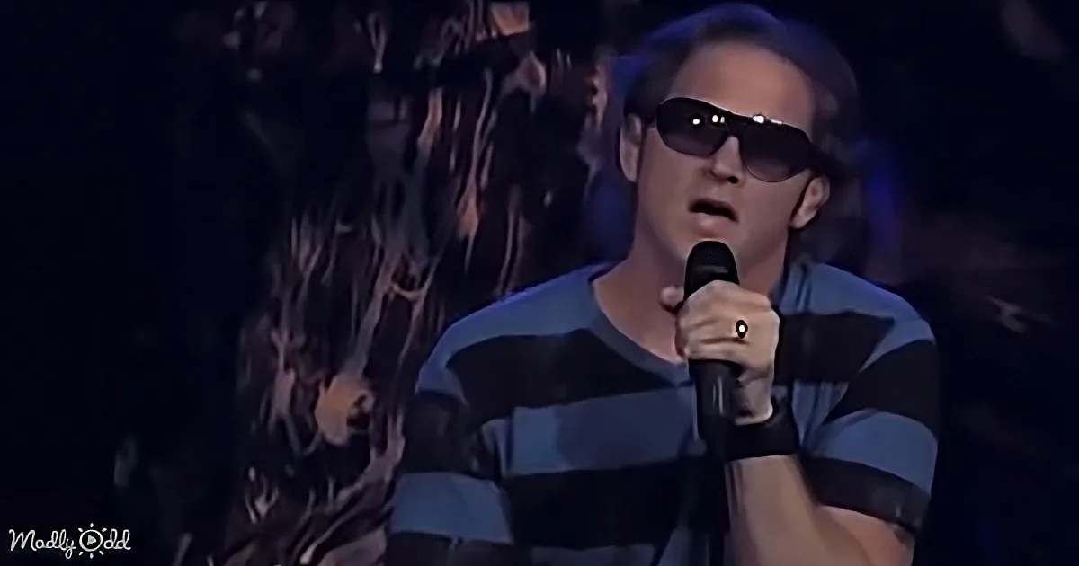 A man in a striped shirt and sunglasses sings into a microphone on stage.