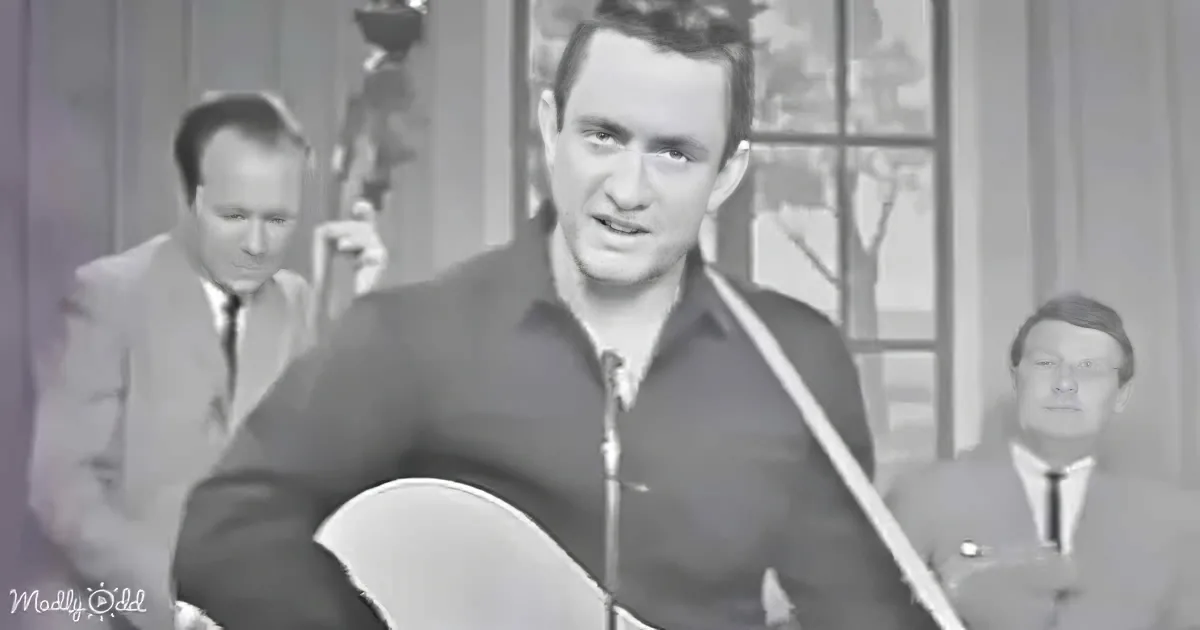 A man in a dark shirt holds a guitar, singing, with a band in the background.