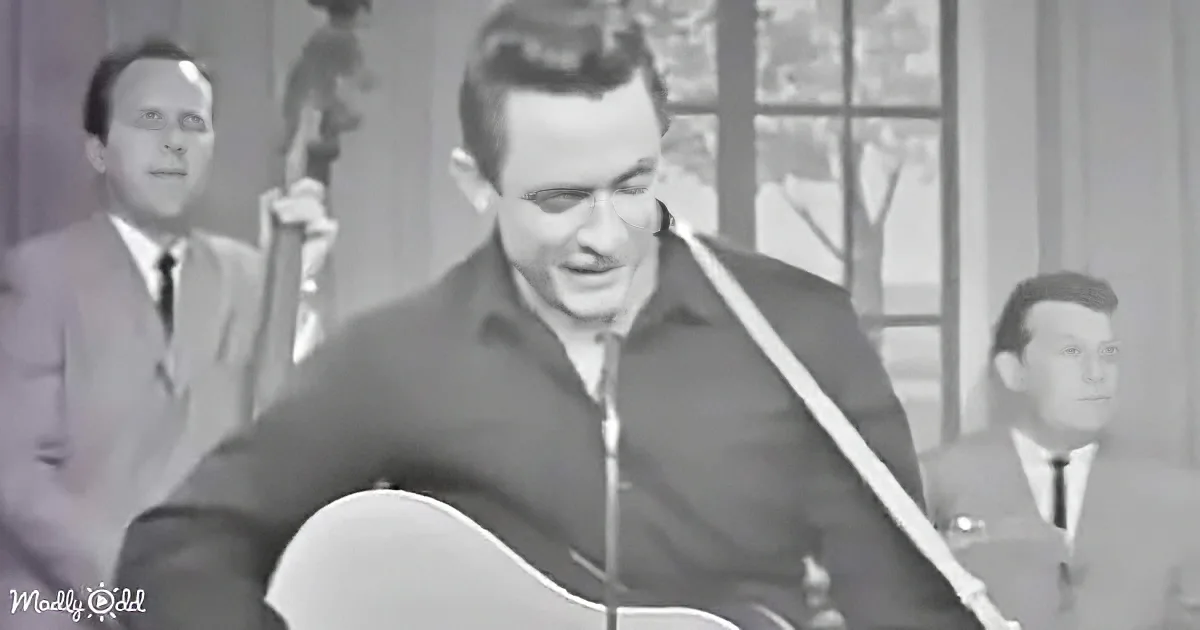 A man in a dark shirt plays guitar in front of a microphone, with another man in a suit and tie to his left.