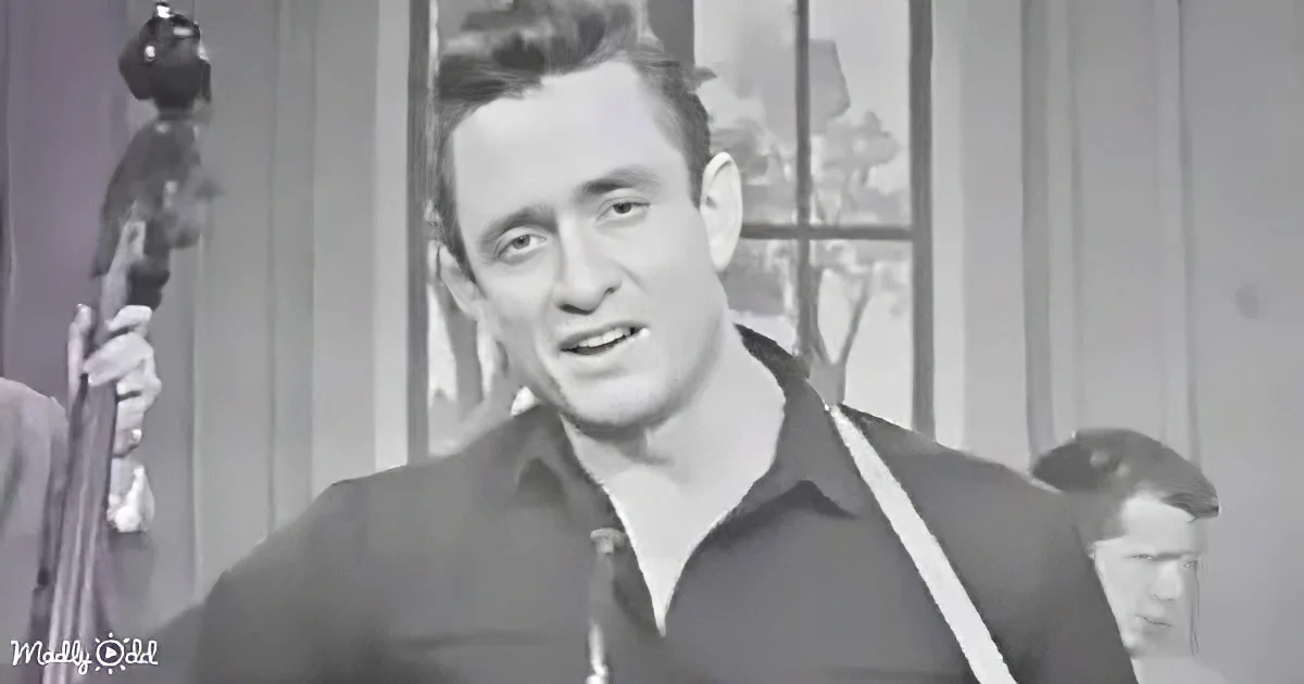 A man in a dark shirt with suspenders is singing, with a band and window in the background.