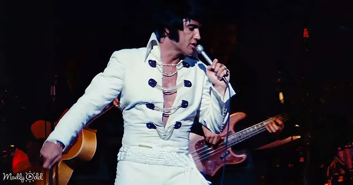 Elvis Presley in white suit singing into microphone, holding guitar, stage performance.