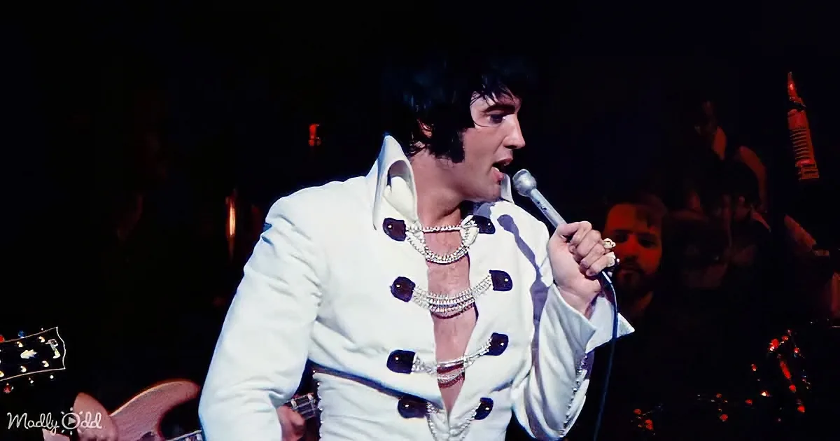 A performer in a white suit with black buttons and a pearl necklace sings into a microphone on stage.