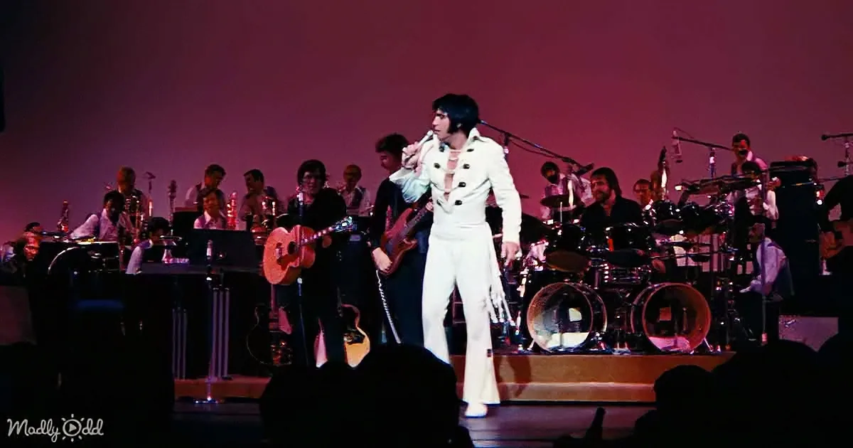 A performer in a white suit sings on stage with a band, surrounded by musicians and instruments.