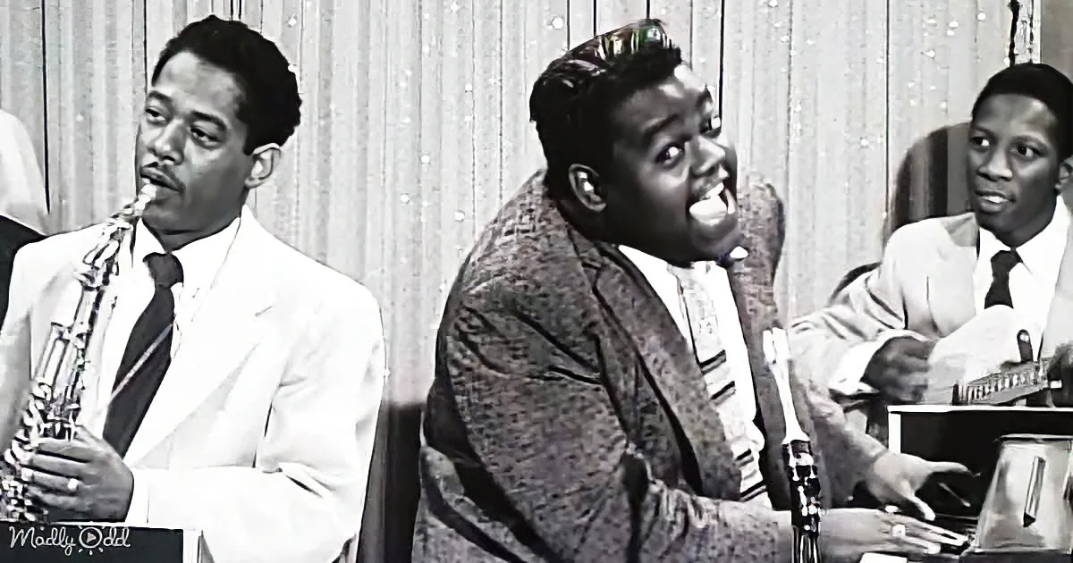 A man in a patterned suit sits at a piano, singing passionately, with a saxophonist and guitarist in the background.