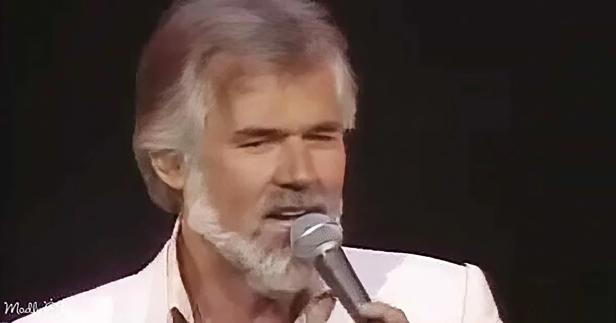 A man with white hair and beard sings into a microphone, wearing a white shirt.