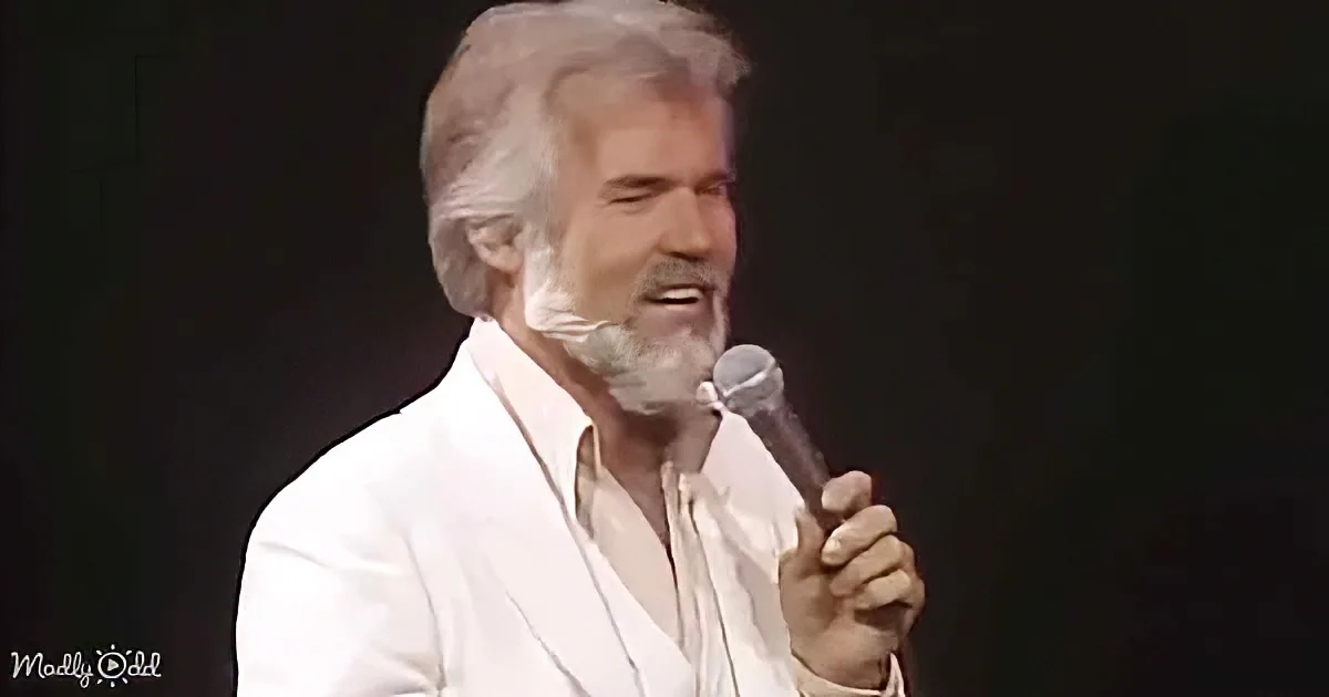 A man with white hair and beard, wearing a white suit, holds a microphone while singing on stage.