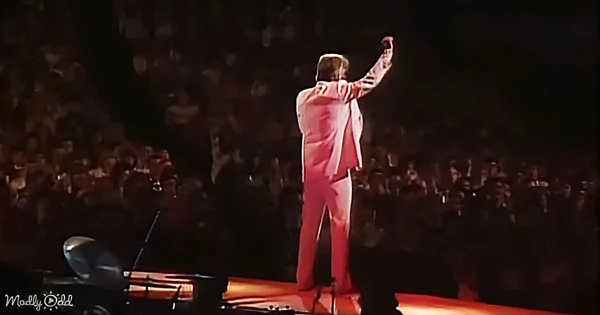 A man in a white suit raises his arm on stage, surrounded by a dark audience.