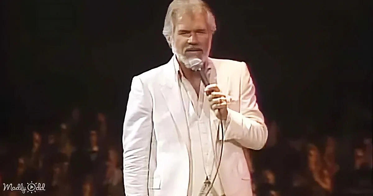 A man in a white suit sings into a microphone on stage, with a blurred audience in the background.