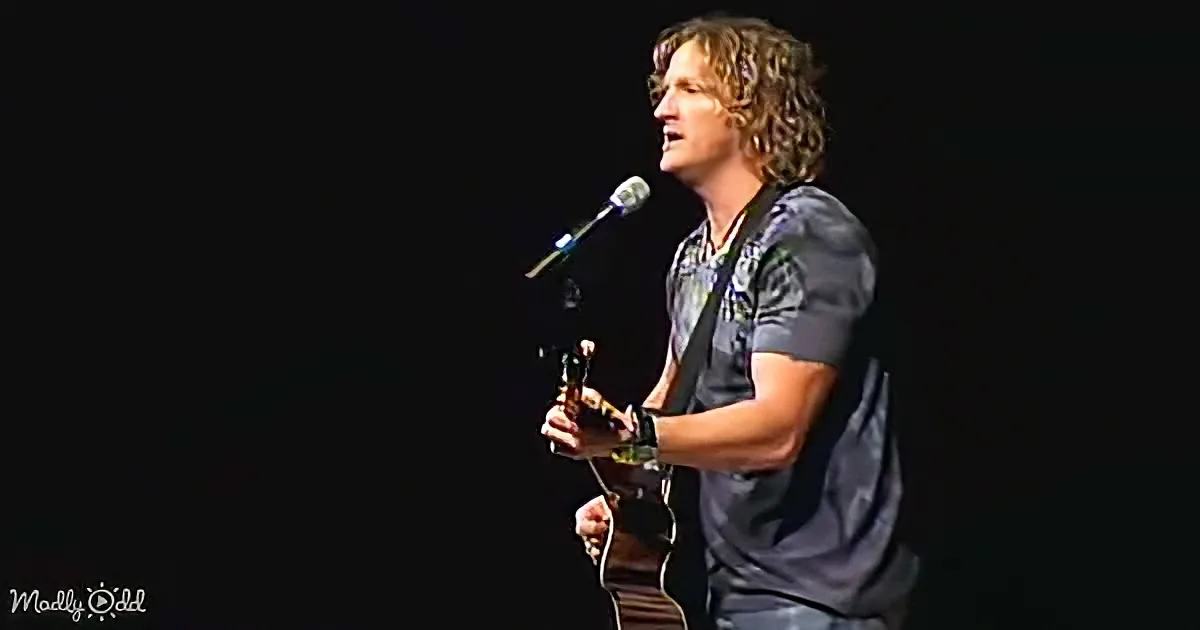 A man with curly hair plays guitar on stage, singing into a microphone.
