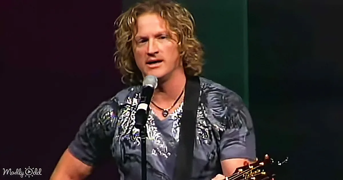 A man with curly hair, wearing a patterned shirt, holds a guitar and sings into a microphone on stage.