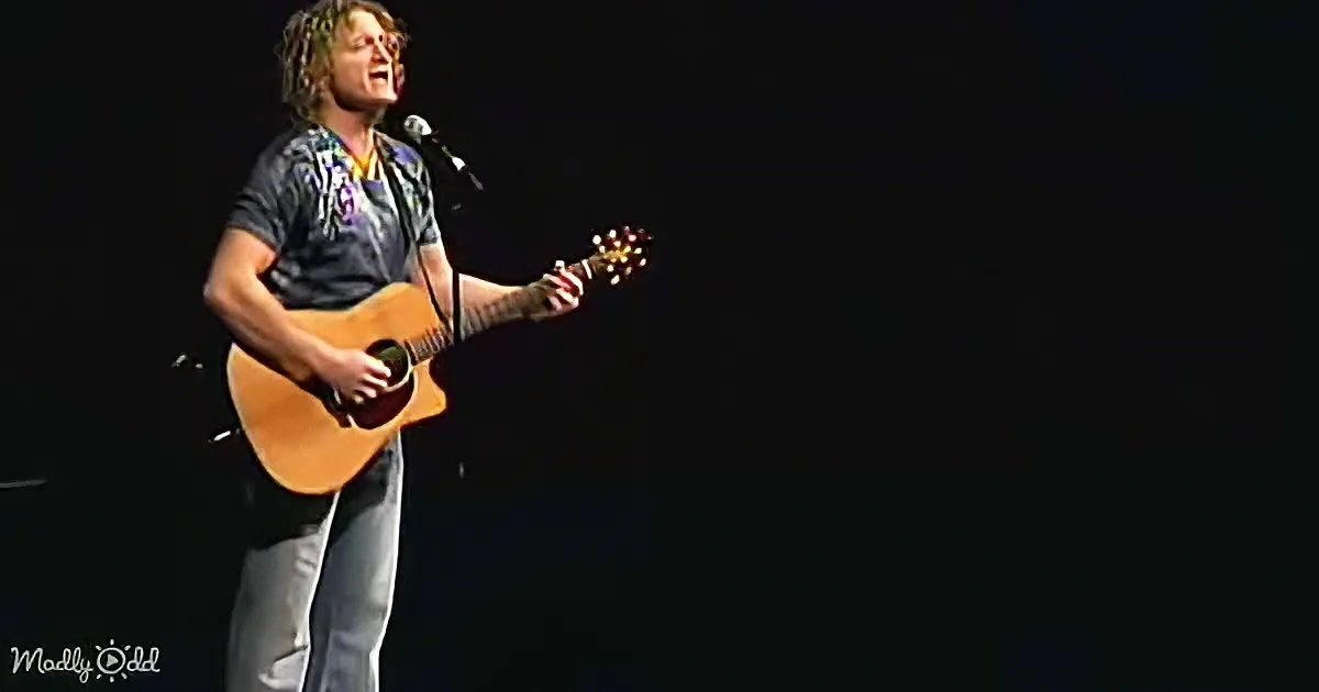 A person with dreadlocks plays an acoustic guitar on stage, singing into a microphone.
