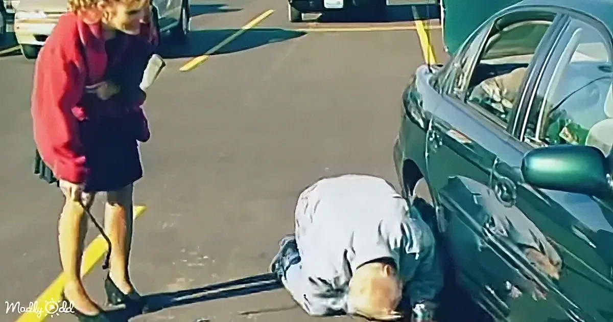 A person in a red jacket stands near a kneeling individual by a car in a parking lot.