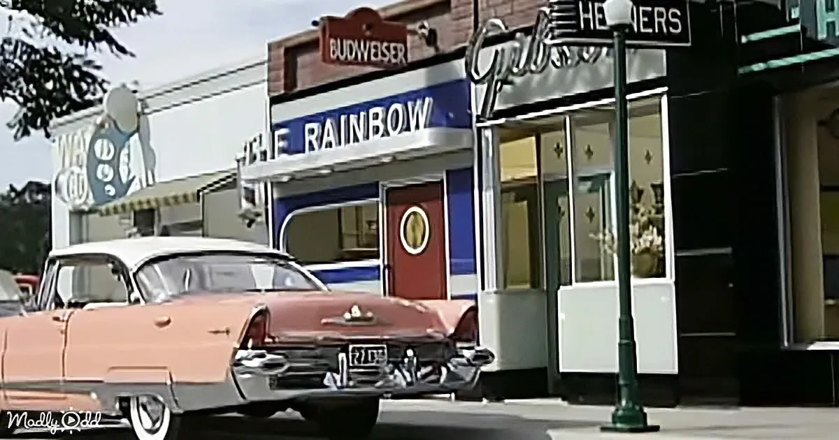 A vintage pink car is parked in front of a "The Rainbow" diner with classic signage and storefronts.