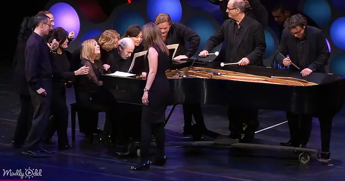 A group of people gathered around a grand piano on stage, some holding mallets, others in conversation.