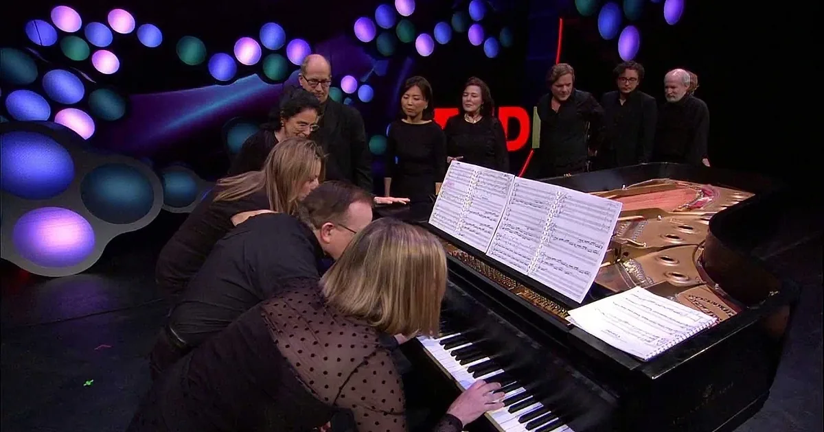 A group of people, including musicians, gather around a grand piano on a stage.