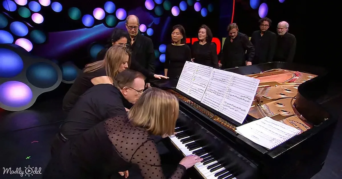 A group of people, including a pianist, are gathered around a grand piano with sheet music, preparing for a performance.