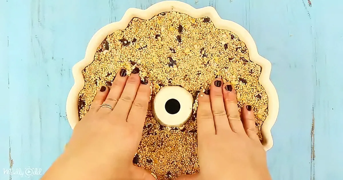 Two hands press down on birdseed mixture in a white dish against a blue wooden background.