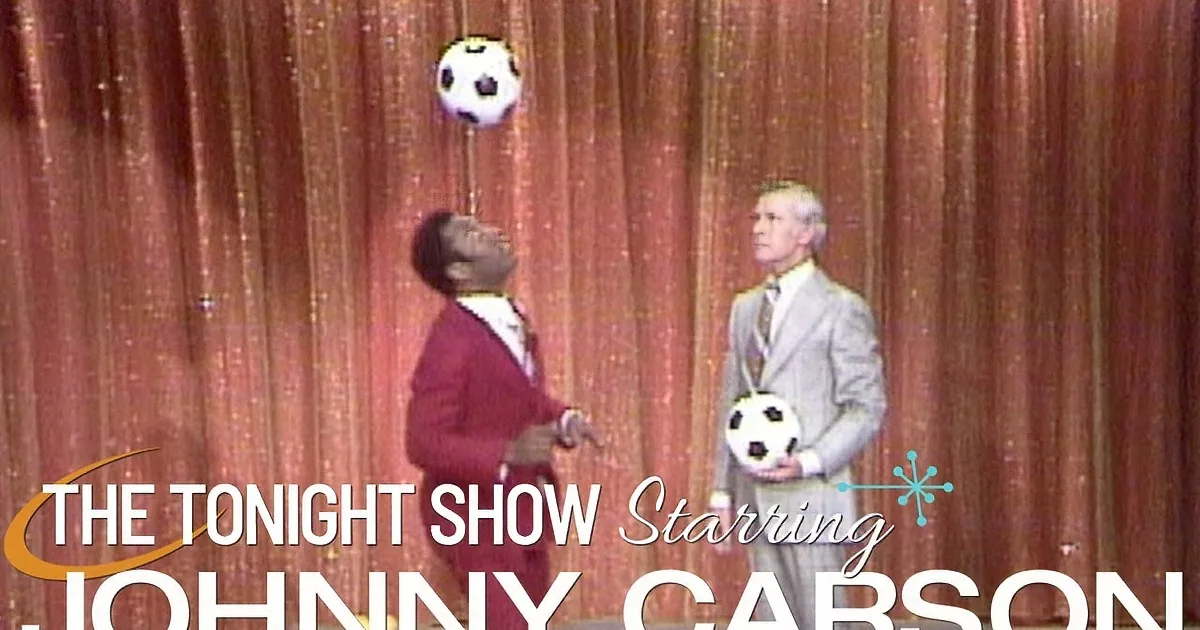 A man in a red suit and polka dot shirt is seated next to a man in a gray suit on a talk show set.