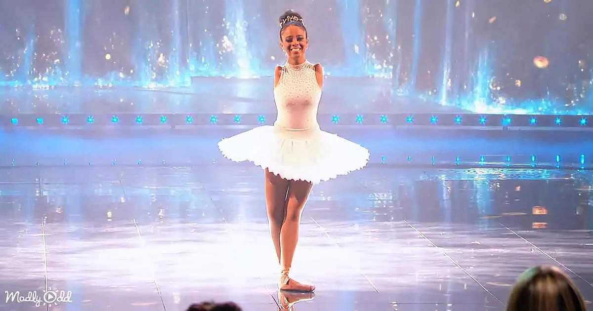 A ballerina in a white tutu smiles on stage with blue lights and water fountains in the background.