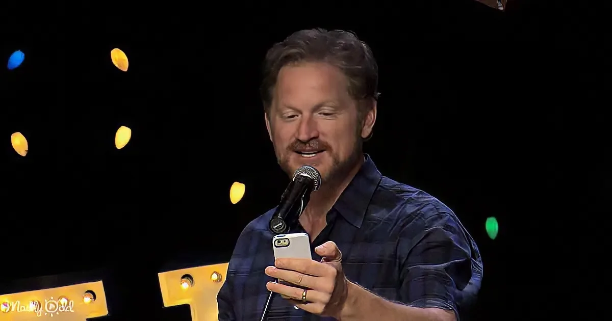 A man in a blue shirt holds a microphone and a phone, speaking on stage with colorful lights in the background.