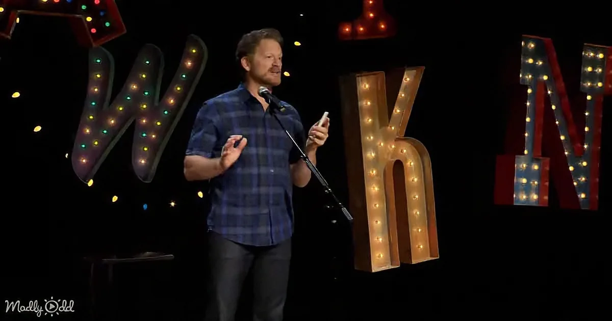A man in a plaid shirt stands on stage with a microphone, gesturing, surrounded by illuminated letters.