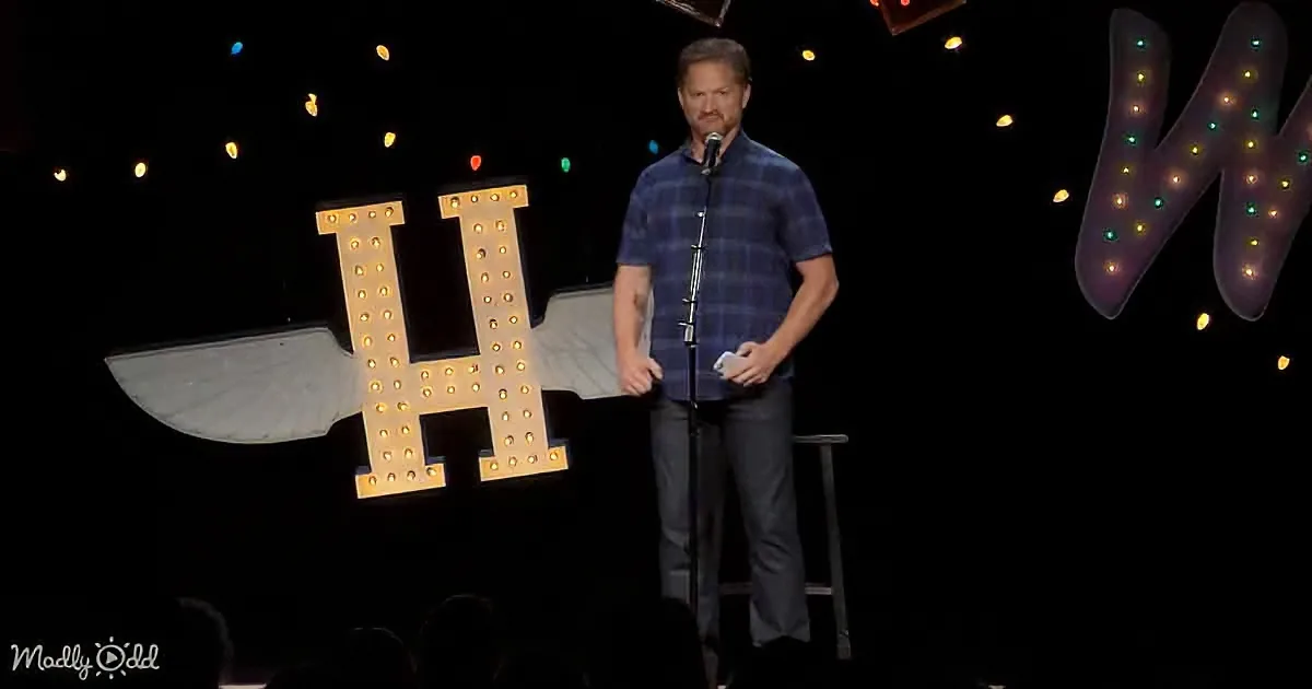 A man in a plaid shirt stands on stage with a microphone, illuminated letters "H" and "K" behind him.