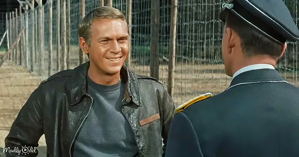 A man in a leather jacket smiles while talking to a uniformed officer in front of a fence.