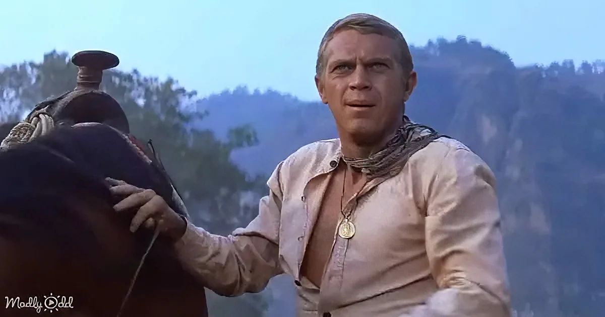 A man in a light shirt and scarf stands beside a horse, with a scenic mountain backdrop.