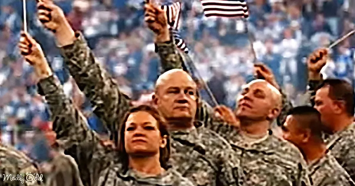 Soldiers in camouflage uniforms raise their hands and hold American flags, standing in a crowd.